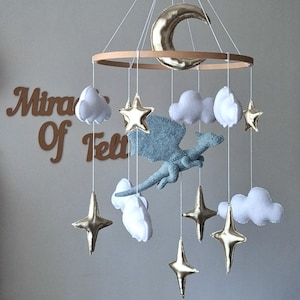 Puede incluir: Un m&oacute;vil colgante que presenta un drag&oacute;n central de fieltro azul claro rodeado de nubes de fieltro blanco, estrellas met&aacute;licas doradas y una luna creciente met&aacute;lica dorada suspendidos de un marco de madera circular con hilos blancos.