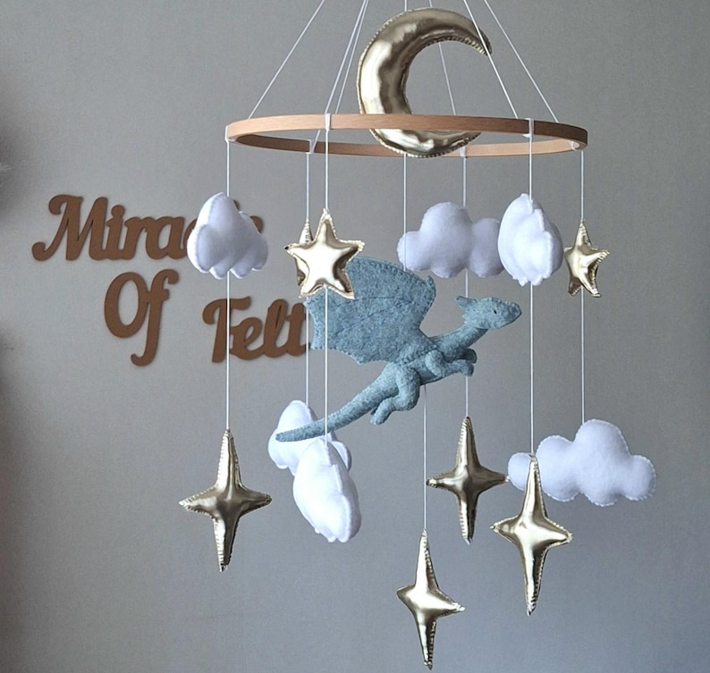 Puede incluir: Un m&oacute;vil colgante que presenta un drag&oacute;n central de fieltro azul claro rodeado de nubes de fieltro blanco, estrellas met&aacute;licas doradas y una luna creciente met&aacute;lica dorada suspendidos de un marco de madera circular con hilos blancos.