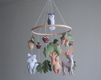 May include: A hanging nursery mobile featuring a circular wooden frame with felt woodland elements suspended by white strings. The felt figures include a grey owl, a grey fox, a brown bear, a tan deer, green oak leaves, brown acorns, and pinecones.