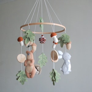 May include: A hanging nursery mobile featuring a circular wooden frame with white strings suspending felt figures of a brown bear, a gray rabbit, a hedgehog, mushrooms, acorns, a pinecone, and green oak leaves in a woodland-themed arrangement.