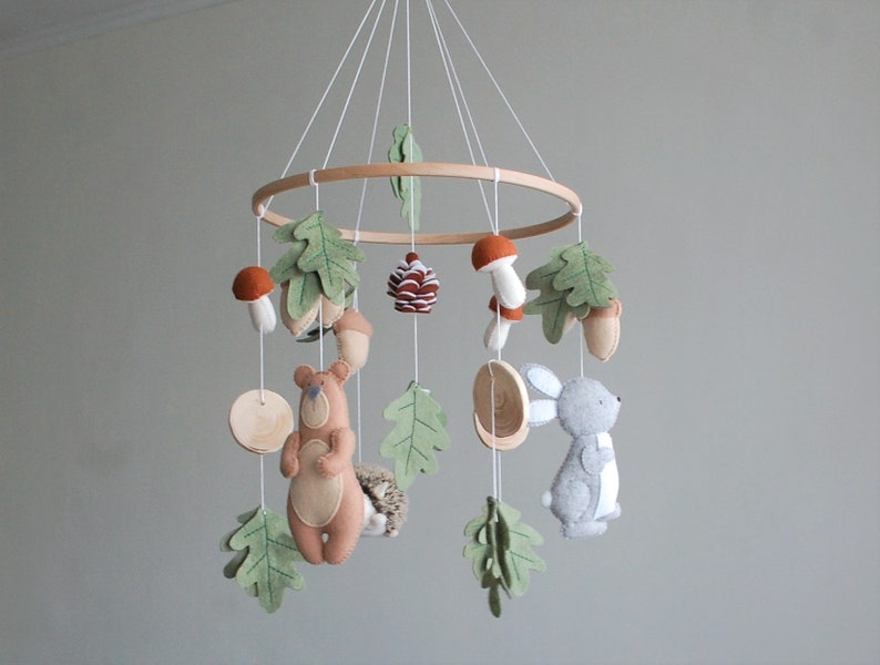 May include: A hanging nursery mobile featuring a circular wooden frame with white strings suspending felt figures of a brown bear, a gray rabbit, a hedgehog, mushrooms, acorns, a pinecone, and green oak leaves in a woodland-themed arrangement.