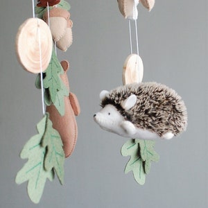 May include: A hanging mobile featuring a plush hedgehog with a brown and white textured back, felt oak leaves in shades of green, tan felt acorns, a brown felt bear silhouette, a brown pinecone, and circular wooden discs suspended by white strings.