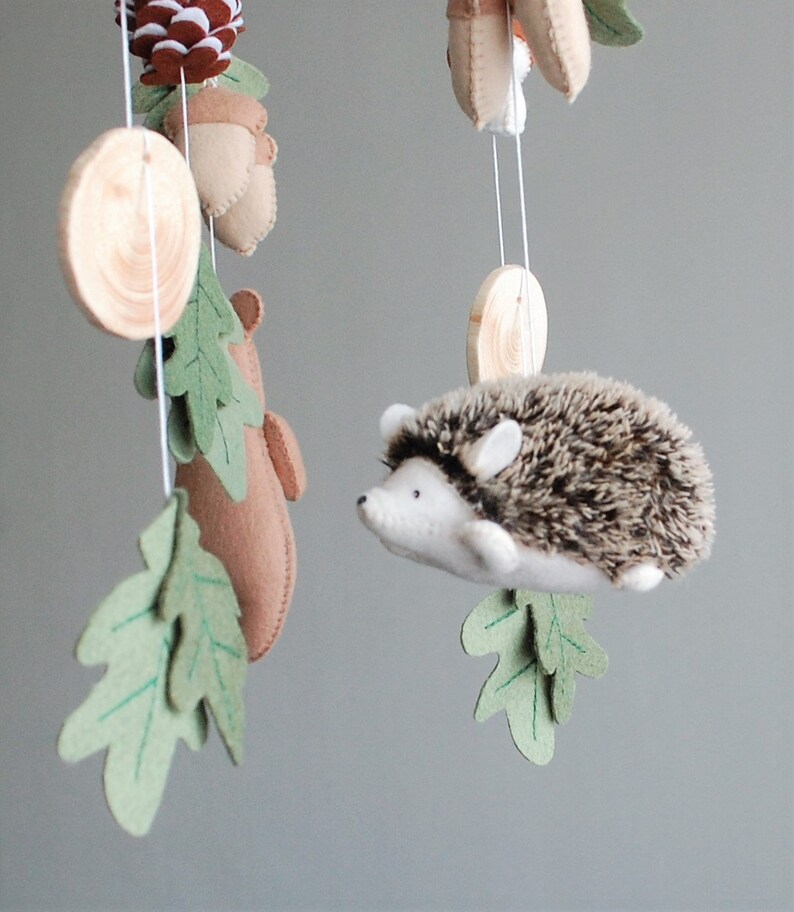 May include: A hanging mobile featuring a plush hedgehog with a brown and white textured back, felt oak leaves in shades of green, tan felt acorns, a brown felt bear silhouette, a brown pinecone, and circular wooden discs suspended by white strings.
