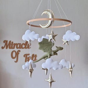 Pode incluir: Um m&oacute;bile suspenso com um drag&atilde;o central de feltro verde-oliva rodeado por nuvens de feltro branco e estrelas e uma lua crescente em dourado met&aacute;lico, suspensos por uma estrutura circular de madeira. O texto Miracle Of Felt &eacute; vis&iacute;vel ao fundo.