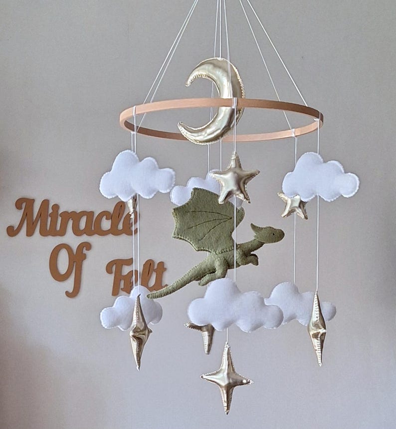 Pode incluir: Um m&oacute;bile suspenso com um drag&atilde;o central de feltro verde-oliva rodeado por nuvens de feltro branco e estrelas e uma lua crescente em dourado met&aacute;lico, suspensos por uma estrutura circular de madeira. O texto Miracle Of Felt &eacute; vis&iacute;vel ao fundo.