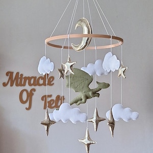 Pode incluir: Um m&oacute;bile suspenso com um drag&atilde;o central de feltro verde-oliva rodeado por nuvens de feltro branco e estrelas e uma lua crescente em dourado met&aacute;lico, suspensos por uma estrutura circular de madeira. O texto Miracle Of Felt &eacute; vis&iacute;vel ao fundo.