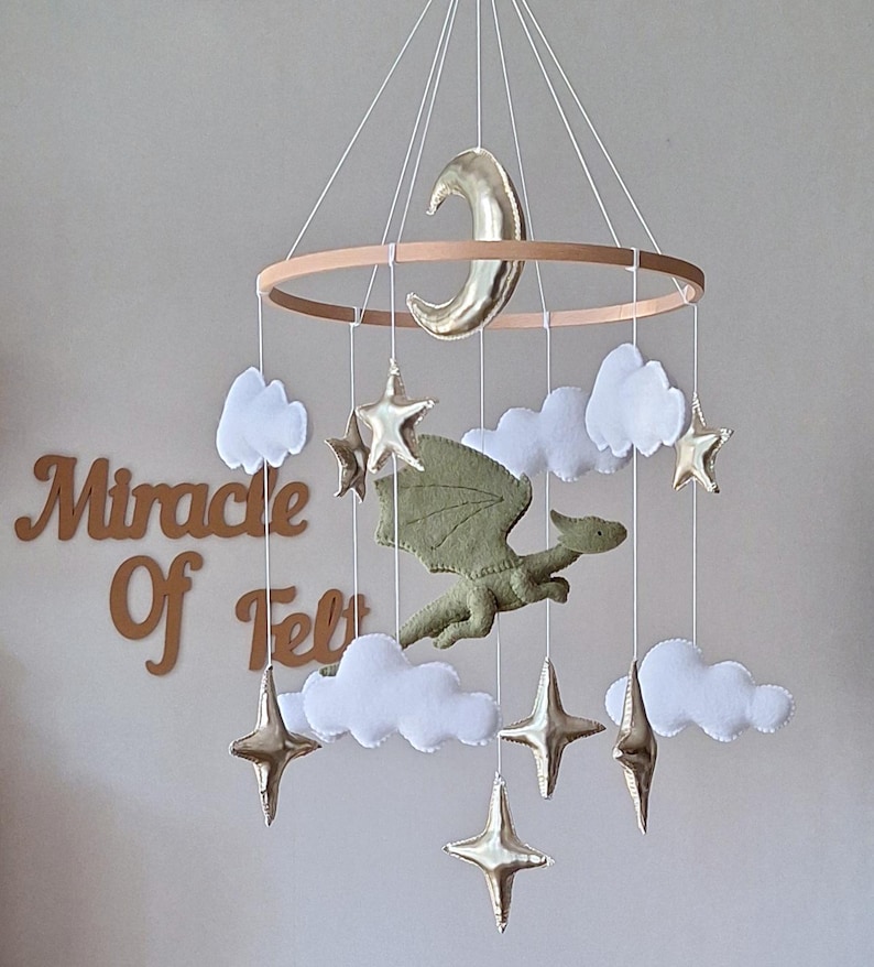 Pode incluir: Um m&oacute;bile suspenso com um drag&atilde;o central de feltro verde-oliva rodeado por nuvens de feltro branco e estrelas e uma lua crescente em dourado met&aacute;lico, suspensos por uma estrutura circular de madeira. O texto Miracle Of Felt &eacute; vis&iacute;vel ao fundo.