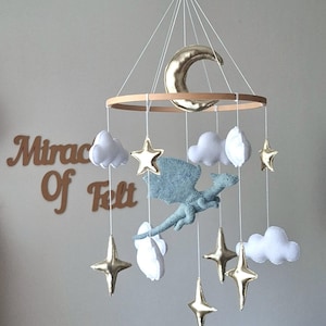 Puede incluir: Un m&oacute;vil colgante para habitaci&oacute;n infantil con un drag&oacute;n central de fieltro azul claro, rodeado de nubes de fieltro blanco y estrellas y una luna creciente de color dorado met&aacute;lico, suspendidos de un marco circular de madera con hilos blancos.