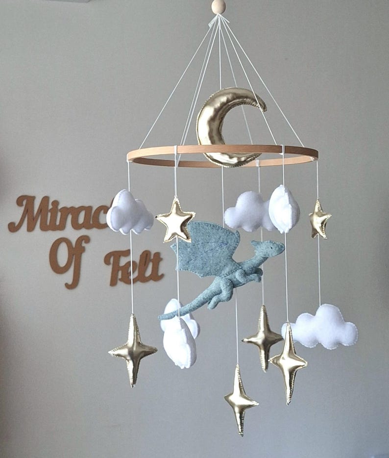 Puede incluir: Un m&oacute;vil colgante para habitaci&oacute;n infantil con un drag&oacute;n central de fieltro azul claro, rodeado de nubes de fieltro blanco y estrellas y una luna creciente de color dorado met&aacute;lico, suspendidos de un marco circular de madera con hilos blancos.