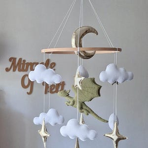 Pode incluir: Um m&oacute;bile suspenso com um drag&atilde;o central de feltro verde-claro rodeado por nuvens de feltro branco, estrelas douradas met&aacute;licas e uma lua crescente, suspensos de uma estrutura circular de madeira por fios brancos finos.