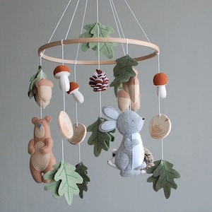May include: A hanging mobile featuring a circular wooden frame with white strings suspending felt figures of a brown bear, a gray rabbit, a hedgehog, orange-capped mushrooms, acorns, a pinecone, and green felt oak leaves with stitched details.