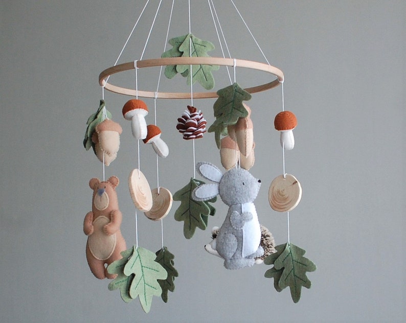 May include: A hanging mobile featuring a circular wooden frame with white strings suspending felt figures of a brown bear, a gray rabbit, a hedgehog, orange-capped mushrooms, acorns, a pinecone, and green felt oak leaves with stitched details.