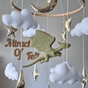 Pode incluir: Um m&oacute;bile suspenso com um drag&atilde;o central de feltro verde s&aacute;lvia, nuvens brancas de feltro, estrelas douradas met&aacute;licas e uma lua crescente suspensas numa estrutura circular de madeira. O texto Miracle Of Felt &eacute; vis&iacute;vel ao fundo.