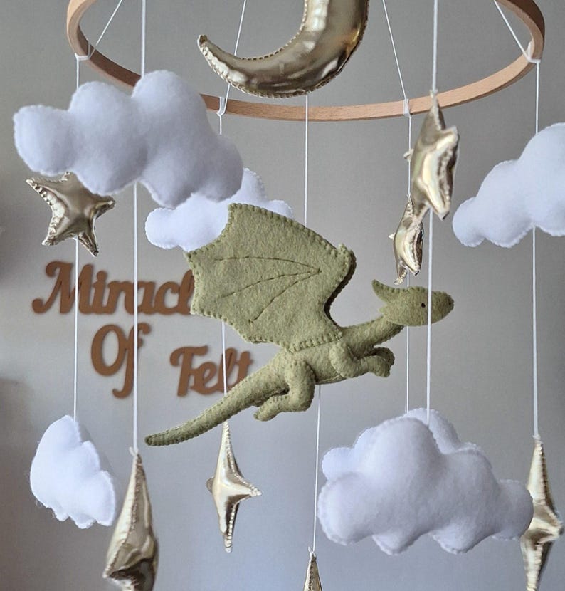 Pode incluir: Um m&oacute;bile suspenso com um drag&atilde;o central de feltro verde s&aacute;lvia, nuvens brancas de feltro, estrelas douradas met&aacute;licas e uma lua crescente suspensas numa estrutura circular de madeira. O texto Miracle Of Felt &eacute; vis&iacute;vel ao fundo.
