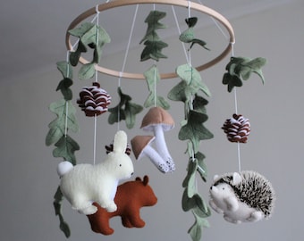 May include: A hanging mobile featuring a circular wooden frame with suspended felt figures including a white rabbit, a brown bear, a hedgehog, two mushrooms, pinecones, and green oak leaves attached to white strings hanging from the hoop.