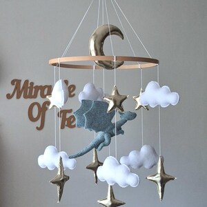Puede incluir: Un m&oacute;vil colgante con un drag&oacute;n central de fieltro gris claro, rodeado de nubes de fieltro blanco y estrellas y una luna creciente de metal dorado, suspendidos de un marco circular de madera con hilos blancos para una exhibici&oacute;n equilibrada.