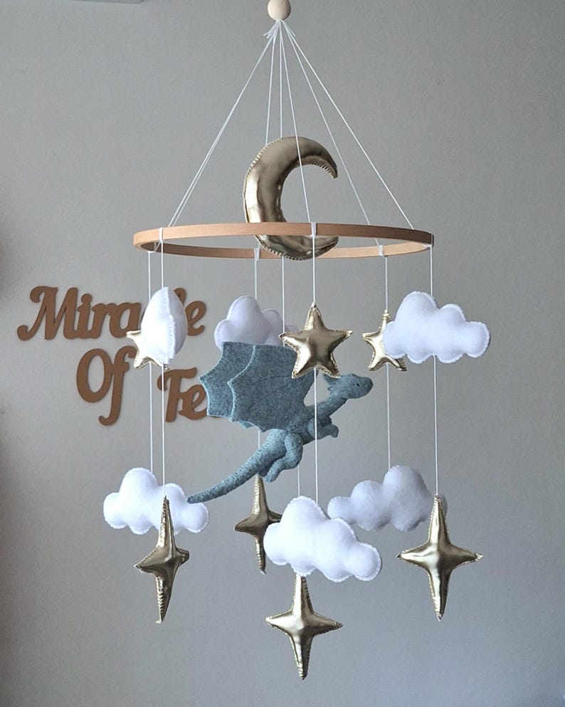 Puede incluir: Un m&oacute;vil colgante con un drag&oacute;n central de fieltro gris claro, rodeado de nubes de fieltro blanco y estrellas y una luna creciente de metal dorado, suspendidos de un marco circular de madera con hilos blancos para una exhibici&oacute;n equilibrada.