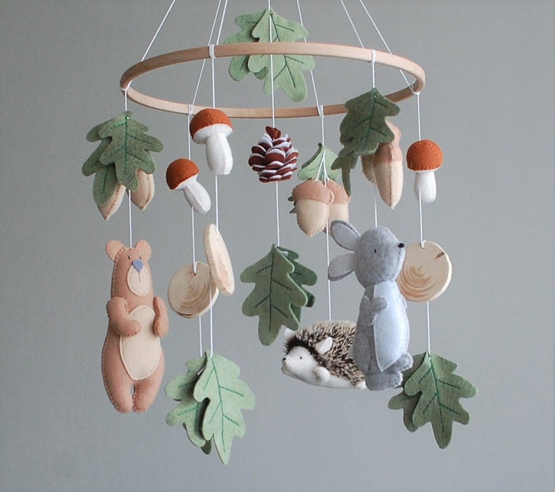 May include: A hanging mobile featuring a circular wooden frame with felt woodland elements including a brown bear, a gray rabbit, a hedgehog, mushrooms, acorns, a pinecone, and green oak leaves suspended by white strings from the wooden ring.