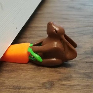 May include: A brown bunny doorstop holding an orange carrot with green leaves. The doorstop is wedged between a white door and a wooden floor. The bunny is in a seated position.