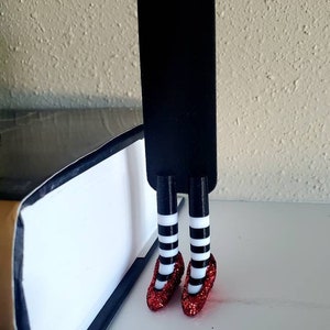 May include: A black bookmark with red glitter shoes and white and black striped stockings. The bookmark is standing upright on a book.