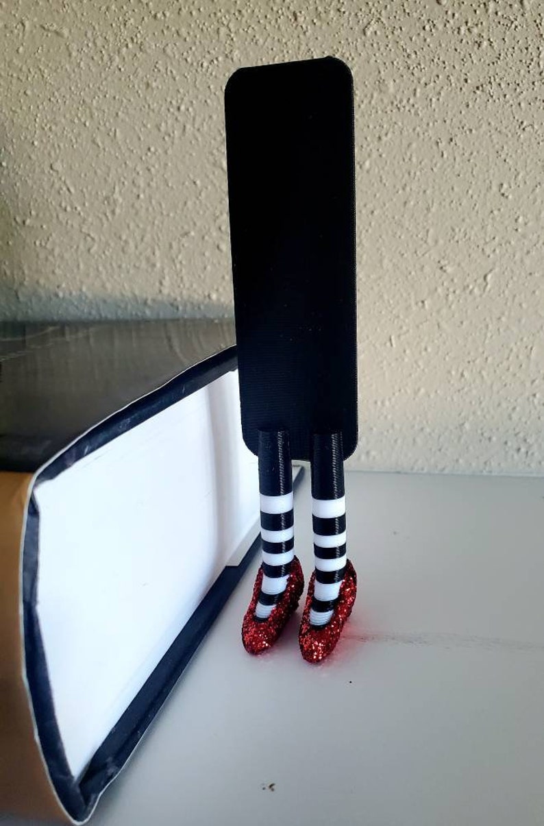 May include: A black bookmark with red glitter shoes and white and black striped stockings. The bookmark is standing upright on a book.
