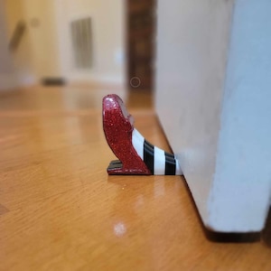 May include: A red glitter shoe with a black and white striped leg doorstop. The shoe is propped against a white door.