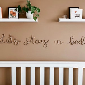 May include: A white wooden headboard with a tan wall behind it. The wall has a rope-like sign that reads "Let's stay in bed".