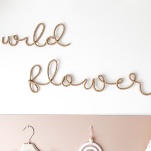 Pu&ograve; includere: Un decoro da parete con le parole "wild flower" realizzate in corda marrone. Le parole sono esposte su una parete bianca.