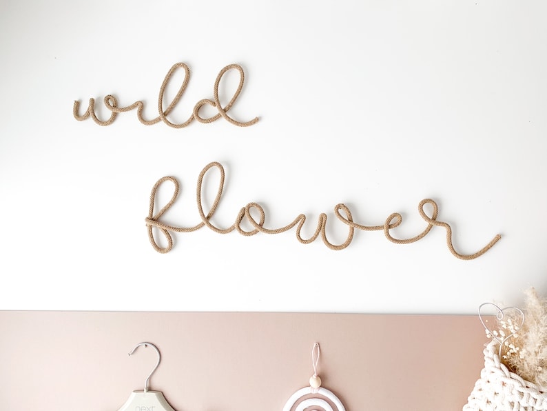 Pu&ograve; includere: Un decoro da parete con le parole "wild flower" realizzate in corda marrone. Le parole sono esposte su una parete bianca.
