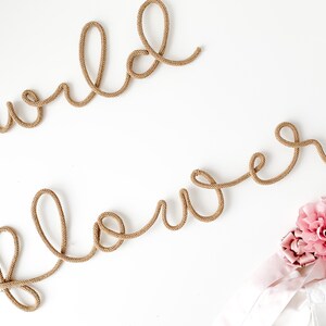 Pu&ograve; includere: Uno sfondo bianco con le parole "wild flower" scritte in corda marrone. Un fiore rosa si trova nell'angolo in basso a destra dell'immagine.