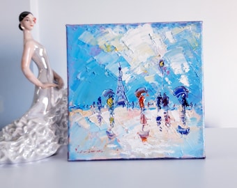 May include: An oil painting on canvas depicting a cityscape with a tall tower in the background. The painting features a blue sky with white clouds, and people walking with umbrellas in the foreground.