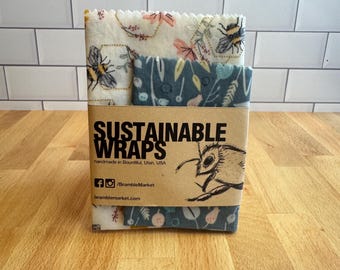 May include: A set of three sustainable food wraps with floral and bee patterns. The wraps are packaged in a brown cardboard sleeve with the words "SUSTAINABLE WRAPS" and a bee illustration. The wraps are handmade in Bountiful, Utah, USA.