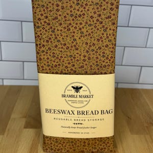 May include: A rectangular beeswax bread bag featuring a brown fabric with a small red and green floral pattern. A cream-colored paper label is wrapped around the center with the text Bramble Market, Beeswax Bread Bag, Reusable Bread Storage, and Handmade in Utah.