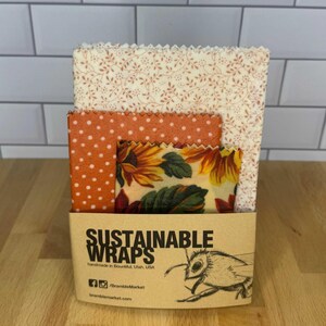 May include: Three rectangular beeswax food wraps with pinked edges, featuring floral and polka dot patterns, held in a tan paper sleeve labeled SUSTAINABLE WRAPS, handmade in Bountiful, Utah, USA, with a bee illustration and social media handles.