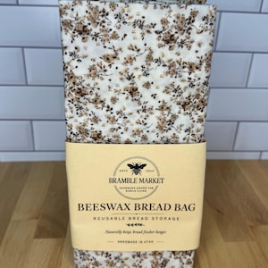 May include: A rectangular beeswax bread bag featuring a white fabric base with a small brown floral pattern. A tan paper label wraps around the center, printed with the text Bramble Market, Beeswax Bread Bag, Reusable Bread Storage, and Handmade in Utah.