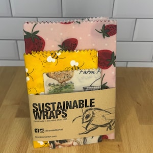 May include: A set of three stacked food wraps with pink strawberry, yellow bee, and cream chicken patterns. A brown paper band wraps around the center, printed with the text SUSTAINABLE WRAPS, handmade in Bountiful, Utah, USA, and a bee illustration.