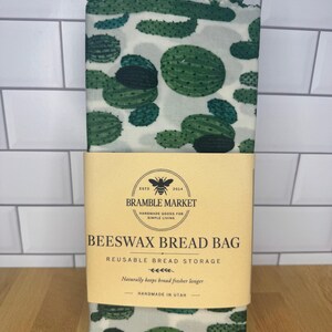 May include: A rectangular beeswax bread bag featuring a green cactus pattern on a light background. A tan paper band wraps around the center with the text BRAMBLE MARKET, ESTD 2014, BEESWAX BREAD BAG, REUSABLE BREAD STORAGE, and HANDMADE IN UTAH.