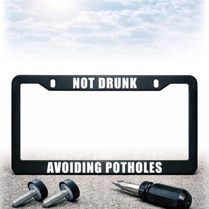 May include: Black license plate frame with the text "NOT DRUNK" at the top and "AVOIDING POTHOLES" at the bottom. Two bolts and a screwdriver are on a concrete surface. The background is a bright sky with clouds.