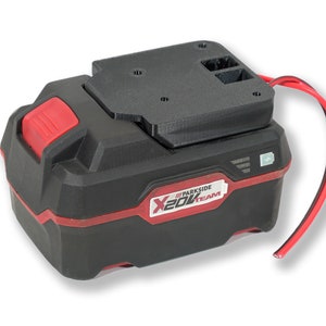 Peut inclure: Une batterie Parkside X20V TEAM noire et rouge avec un adaptateur noir sur le dessus. La batterie a un bouton rouge et une bande rouge. Un fil rouge sort de l'adaptateur. La batterie a un indicateur de puissance.