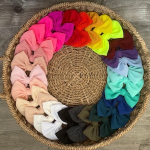 May include: A collection of colorful hair bows arranged in a circle on a woven basket. The bows are made of a soft, textured fabric and come in a variety of colors, including pink, red, orange, yellow, green, blue, purple, and white.