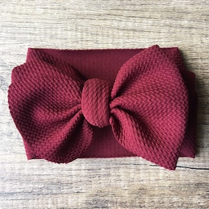 May include: A burgundy fabric headband with a large, knotted bow. The headband is made of a soft, textured fabric.