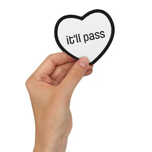 Könnte beinhalten: Ein weißes herzförmiges Abzeichen mit schwarzem Rand, das den Schriftzug "it'll pass" in Schwarz trägt. Das Abzeichen wird in einer Hand vor weißem Hintergrund gehalten.