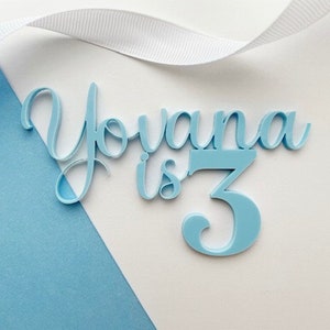 May include: Light blue acrylic cake topper with the name "Yovana is 3" in a cursive font.
