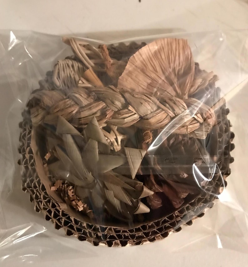 May include: A circular bird toy made from natural, woven plant fibres, presented in a clear plastic bag. It is set within a corrugated cardboard base. The colours are predominantly tan and brown.