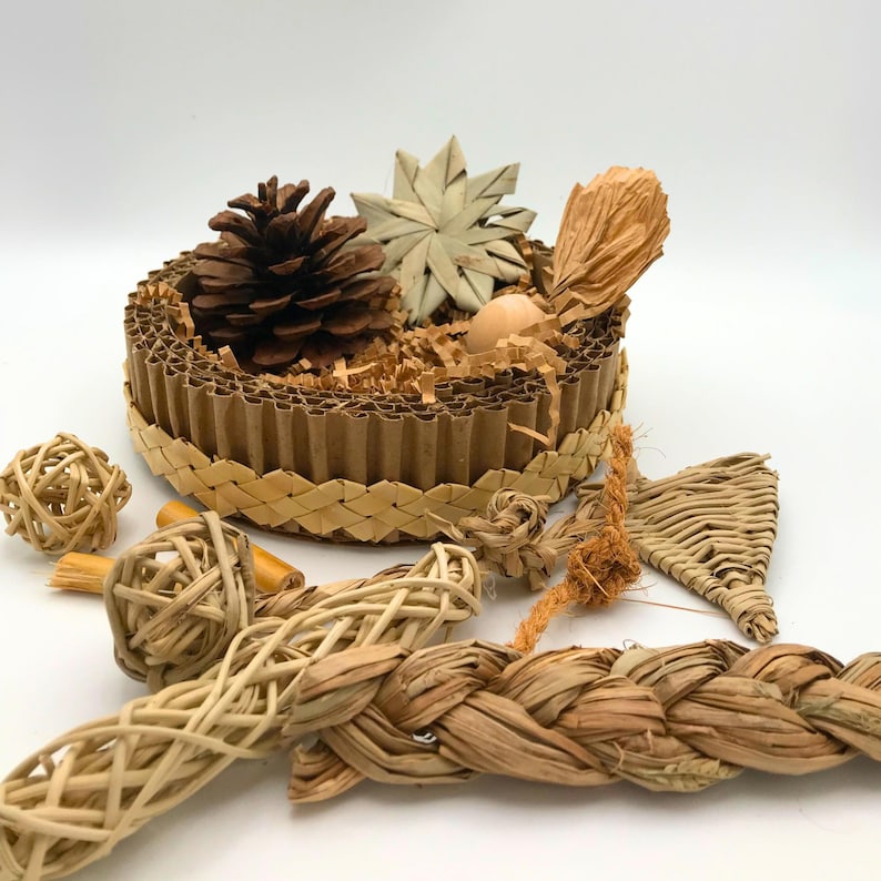 May include: A selection of natural bird toys. A brown cardboard basket contains a pinecone, star-shaped decoration, and shredded paper. Other toys include woven balls, braided rope, and a woven triangle.