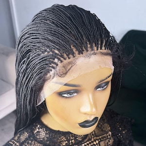 May include: A black braided wig with a lace front is shown on a mannequin head. The wig is made up of many small braids, giving it a textured look. The lace front is visible, and the mannequin has dark makeup, including black lipstick.