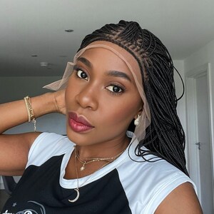 May include: A black braided wig with a lace front. The wig is styled in small, thin braids. The wig is worn by a person with dark skin and a neutral makeup look. The person is wearing a black and white shirt and gold jewelry.