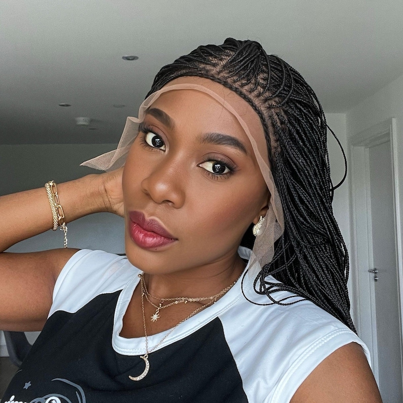 May include: A black braided wig with a lace front. The wig is styled in small, thin braids. The wig is worn by a person with dark skin and a neutral makeup look. The person is wearing a black and white shirt and gold jewelry.