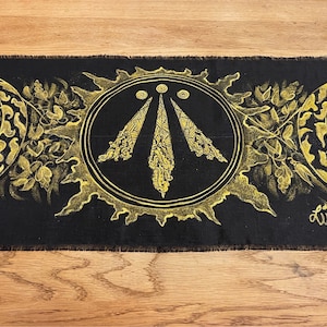 May include: Black fabric banner with a gold sun, moon, and a triple spiral symbol in a circle. The symbol is surrounded by a decorative border. The banner has a frayed edge.