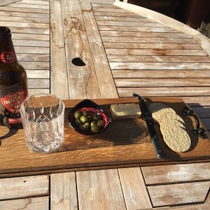May include: A wooden serving tray with a bottle of beer, a glass, a small bowl of olives, cheese, crackers, and a cheese knife. The tray has black metal handles and is set on a wooden table.